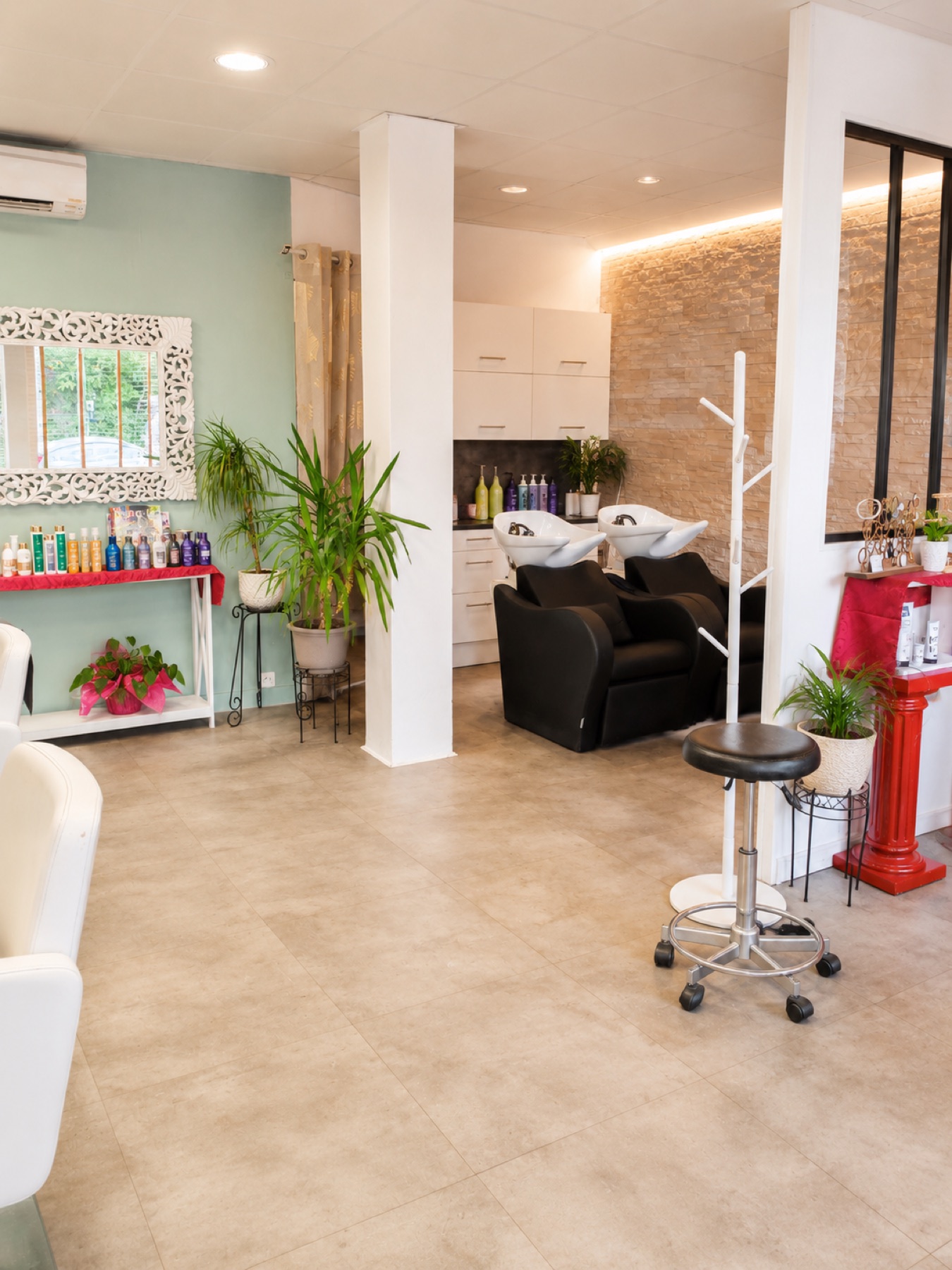 Espace shampoing du salon : bacs à shampoing noirs sous mur de pierre claire, mur vert sauge avec présentoir produits, plantes vertes et présentoir rouge à droite