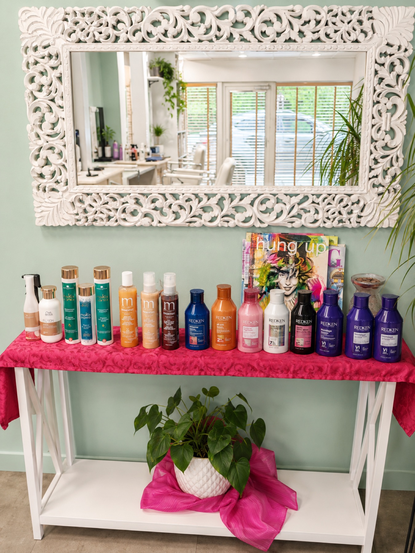 Présentoir produits sous grand miroir blanc finement sculpté, alignement de soins et shampoings professionnels, plante verte et drapé rose, mur vert sauge